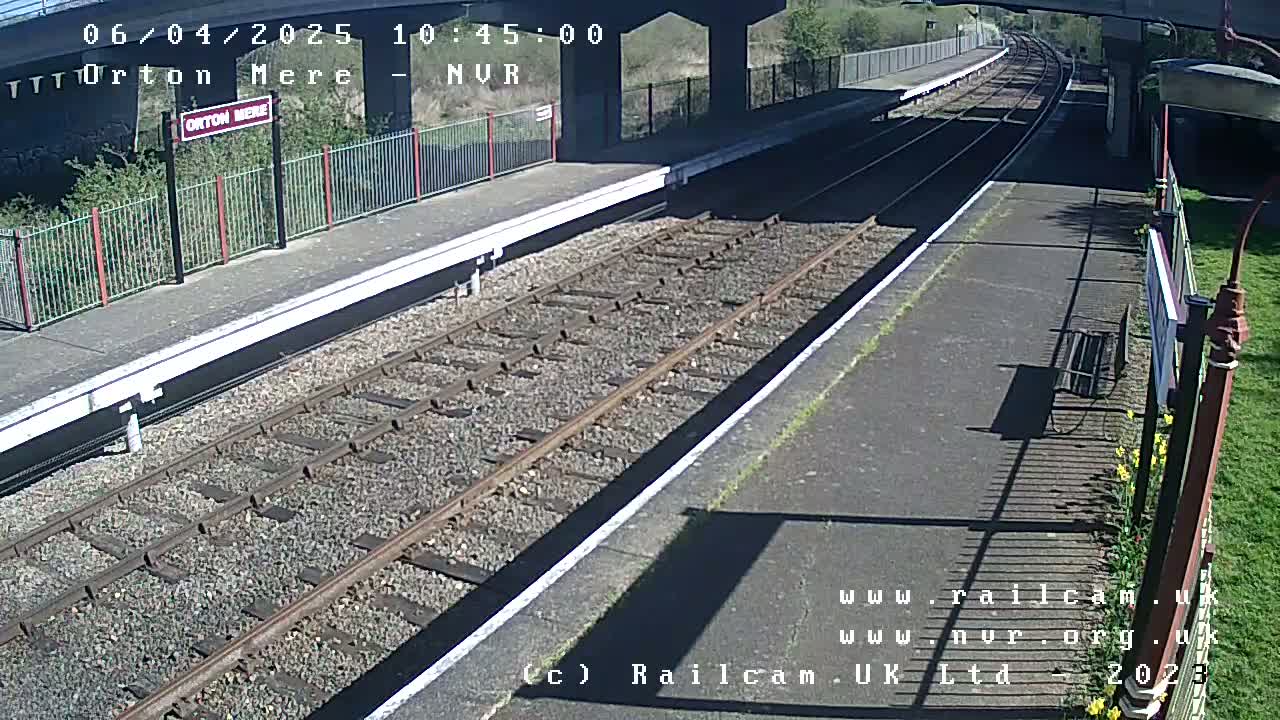 A sunny view of a train station platform with railway tracks extending into the distance, under a bridge.