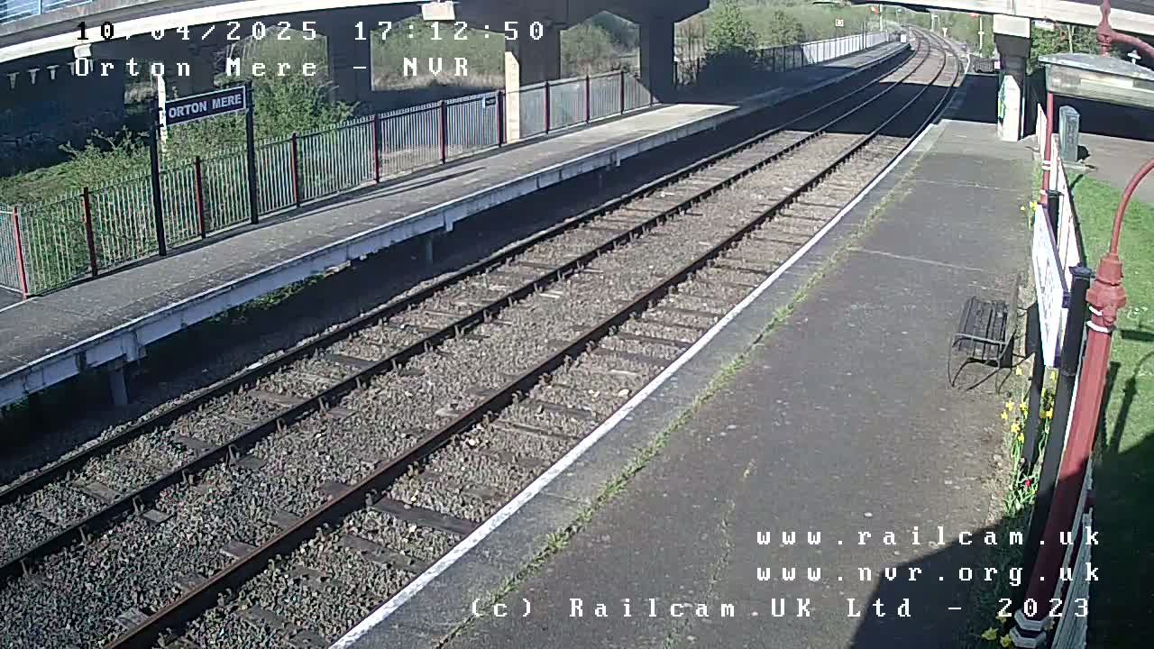 A train platform with railway tracks extending into the distance under a bridge on a sunny day.