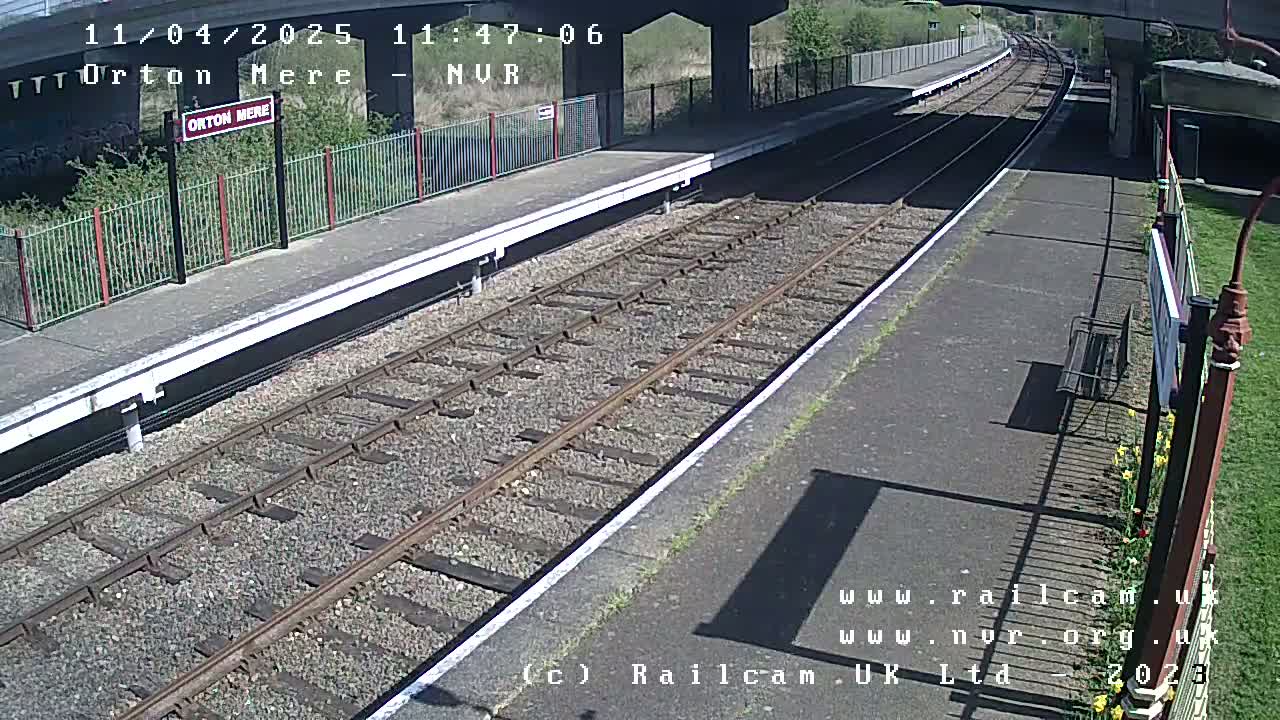 A sunny day at a train station with an empty platform and railway tracks extending into the distance under a bridge.