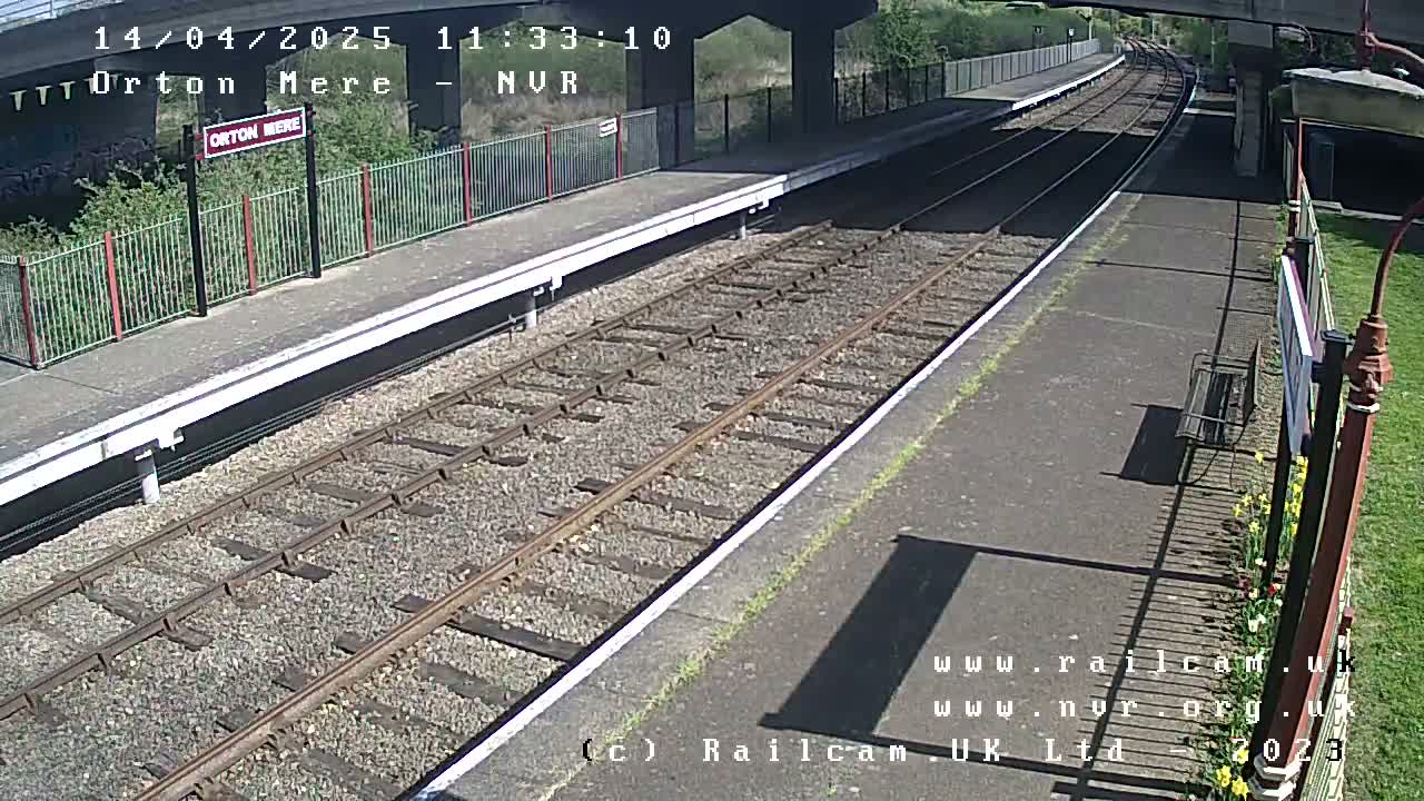 A sunny view of a train platform with railway tracks extending into the distance, under a bridge.