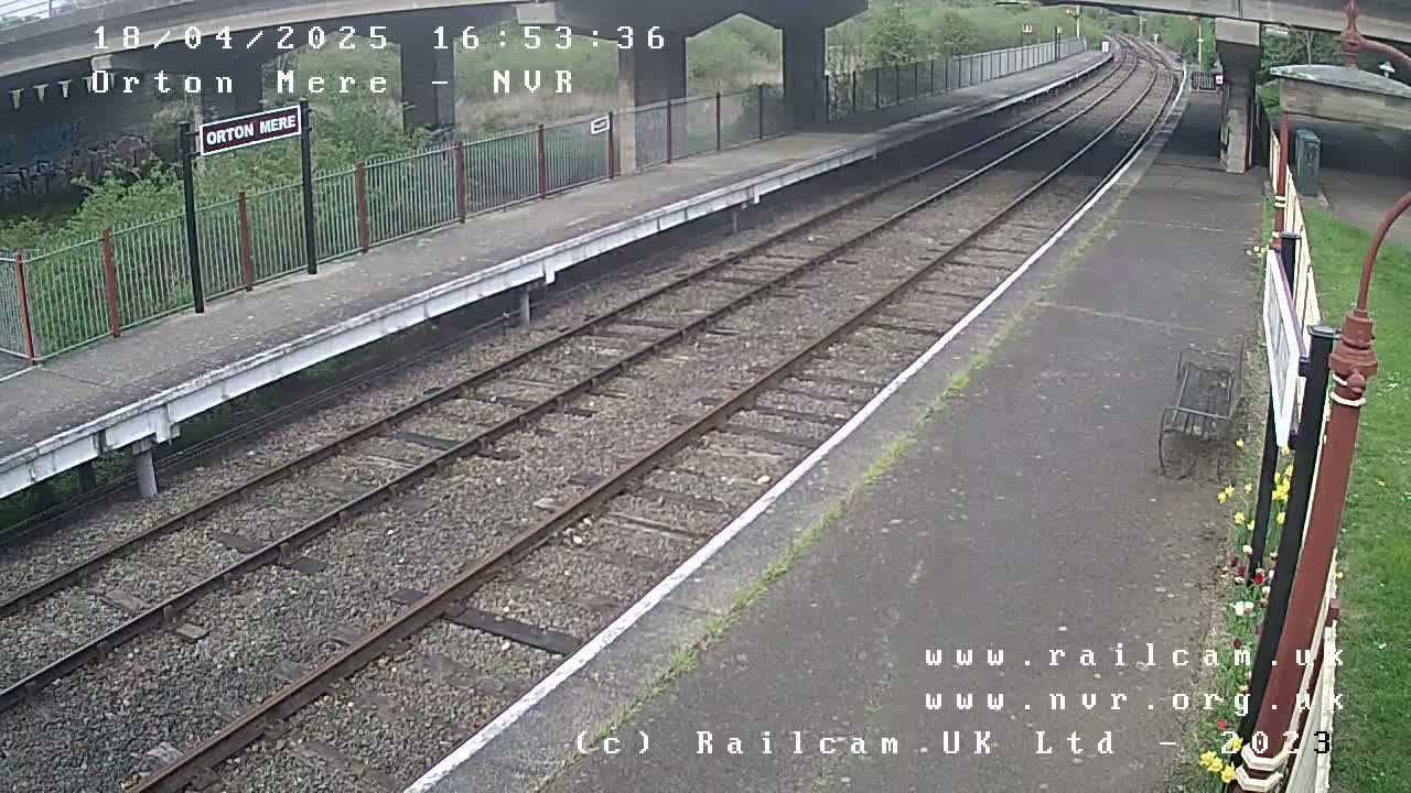 A mostly overcast view of an empty train platform with two sets of train tracks extending into the distance under a bridge.