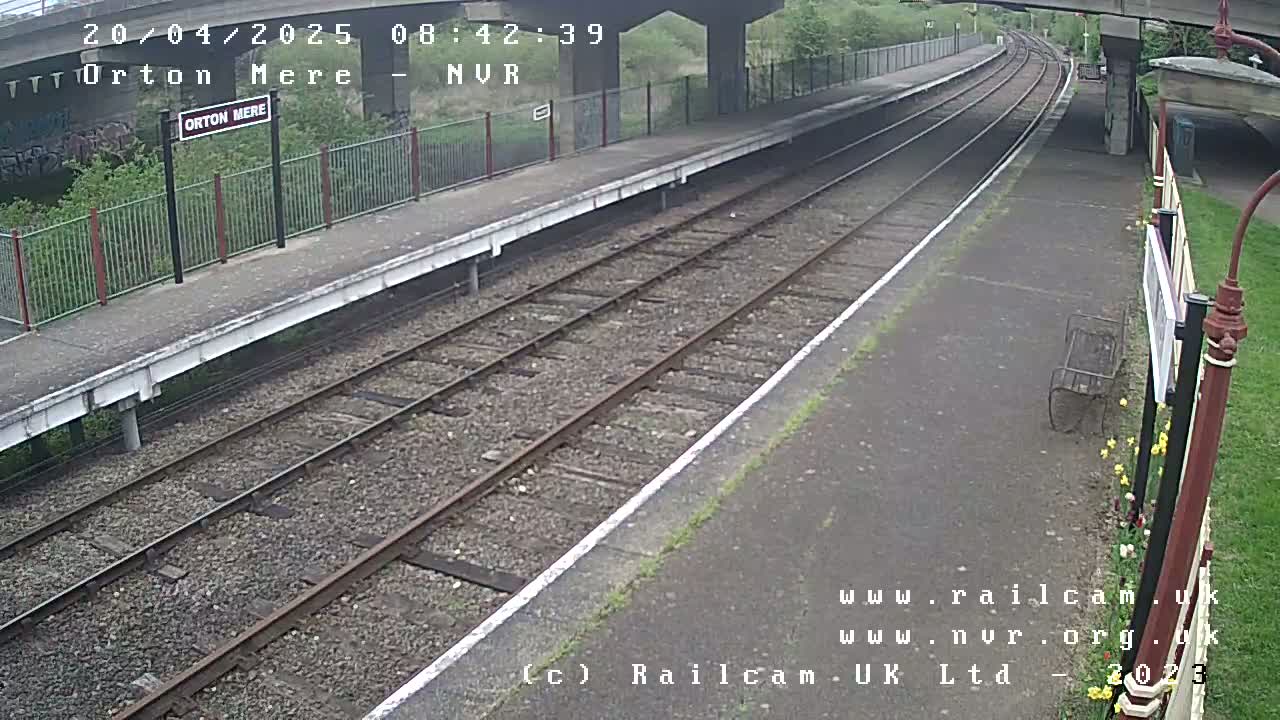 A mostly overcast view of a train platform with two railway tracks and a bench, under a bridge.