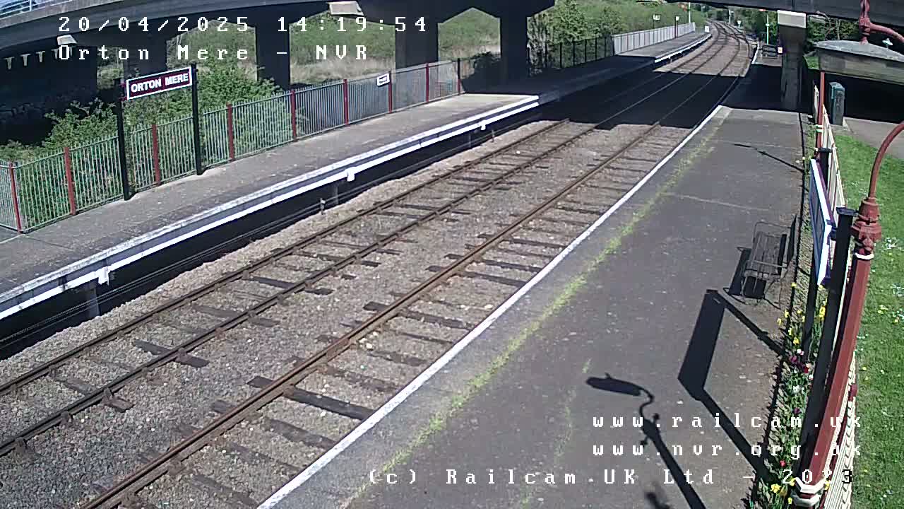 A sunny view of a train station platform with two train tracks extending into the distance, under a bridge.