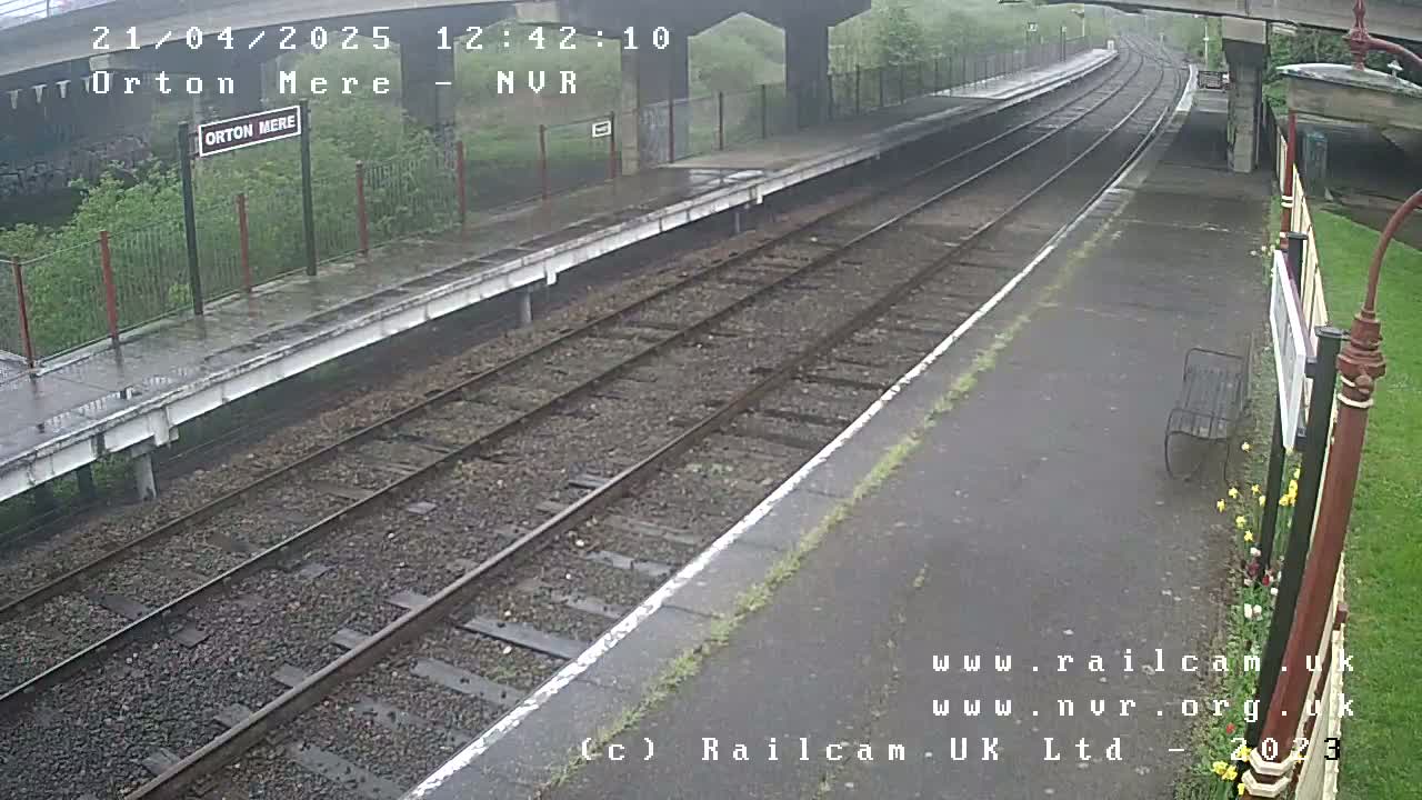 A wet, empty train platform with railway tracks extending into the distance under an overcast sky.