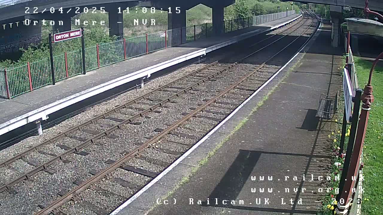 A sunny view of a train platform with two sets of tracks, a bench, and some vegetation.
