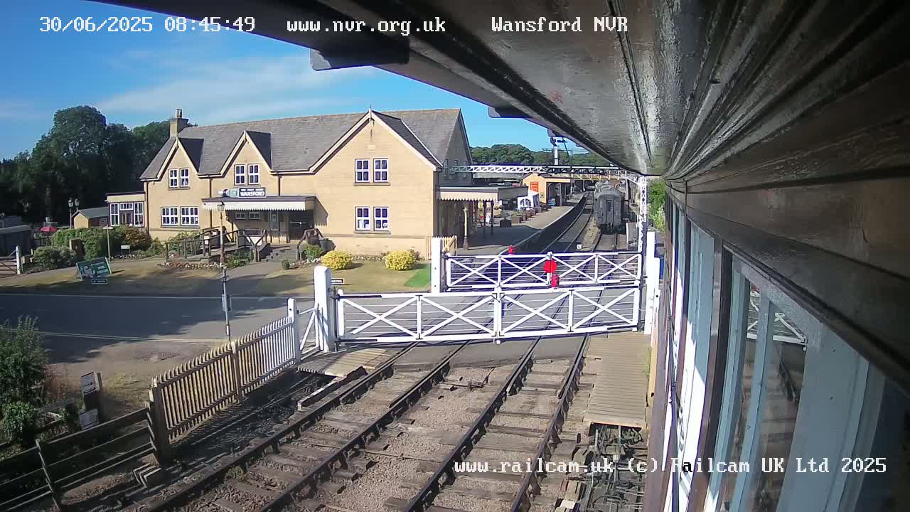 A train approaches a railway station on a sunny day, passing through level crossing gates near a stone building.