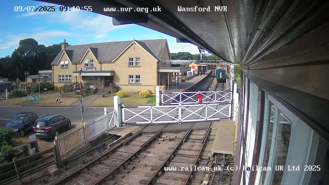 A train passes by a railway station building on a sunny day, with cars parked nearby and a level crossing gate in the foreground.