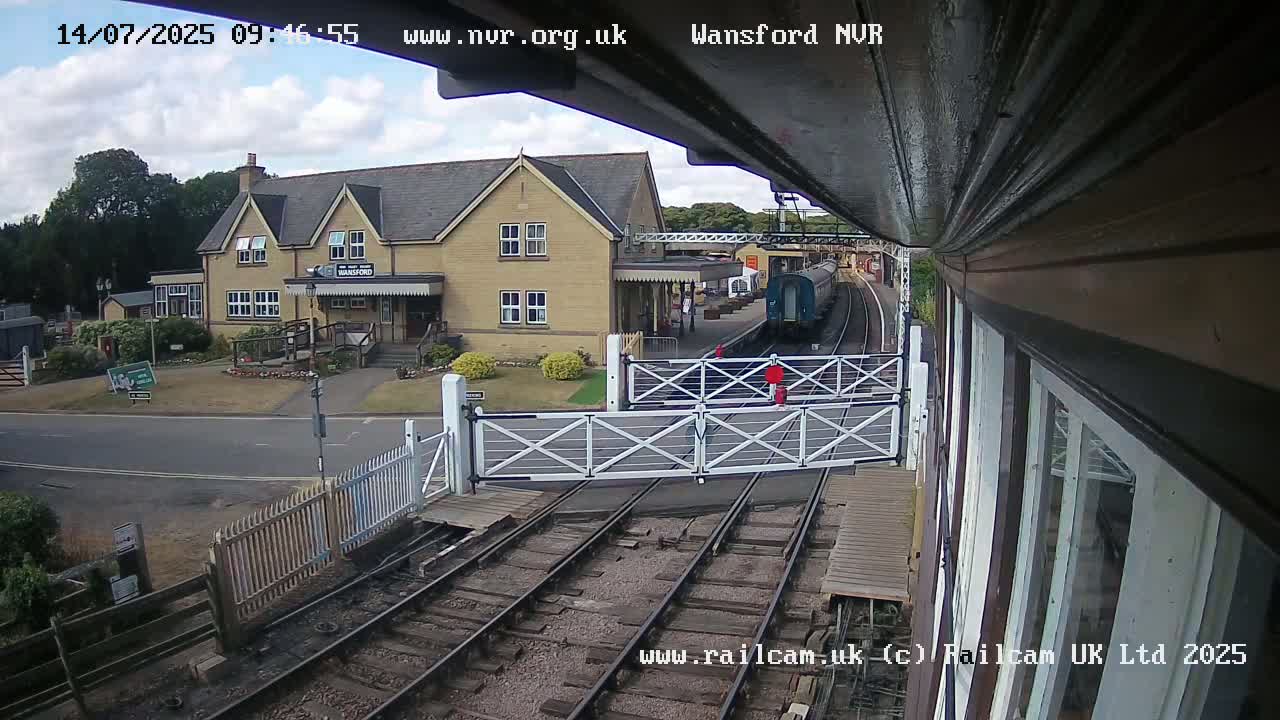 A train is arriving at a station on a partly cloudy day, with the station building and level crossing visible.