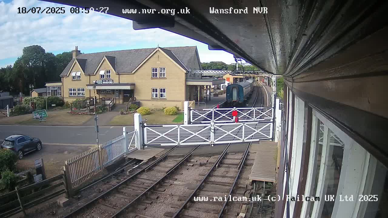 A train is arriving at a station on a partly cloudy day, with crossing gates down and a car parked nearby.