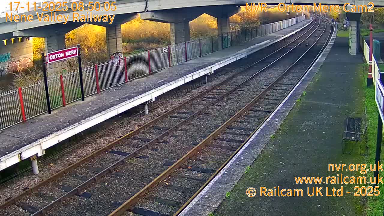 An empty railway station features two platforms alongside multiple tracks that curve into the distance, with a concrete bridge overhead, under bright and clear conditions.