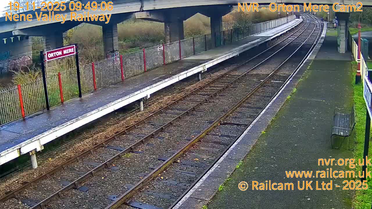 An empty Orton Mere railway station platform and tracks are visible, appearing damp under an overcast sky, with an overhead bridge in the background.
