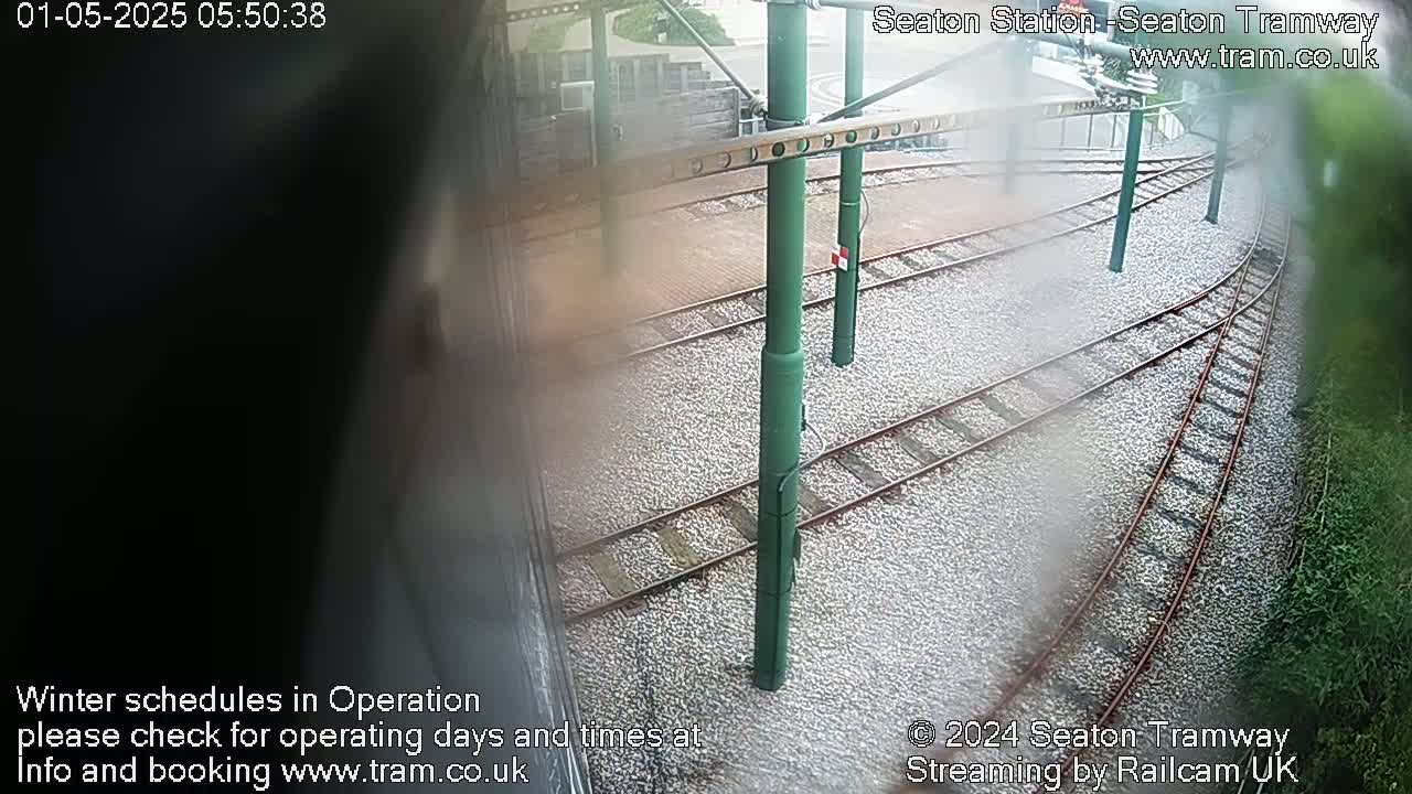 A view from a window shows a section of train tracks in the rain, with green support poles and gravel ballast.