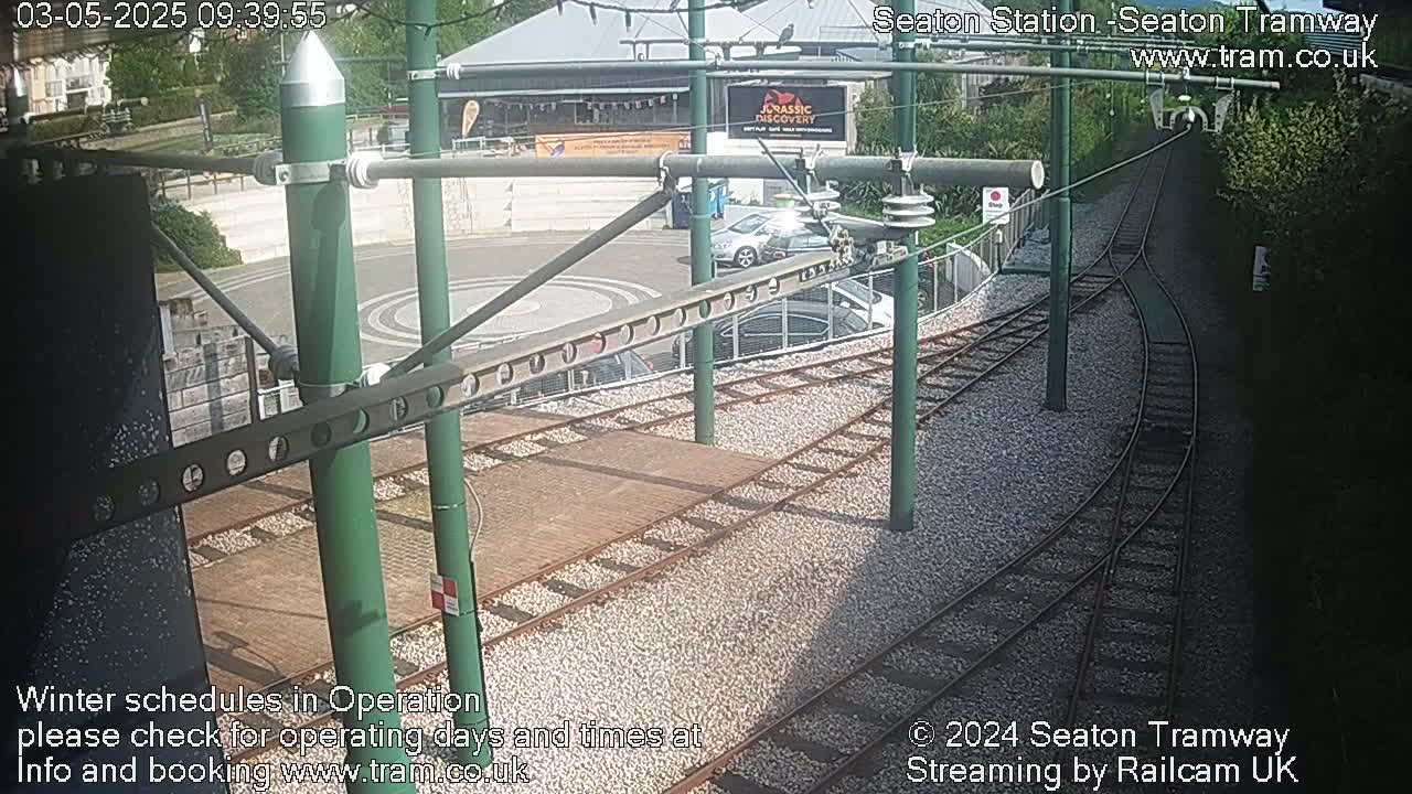 A sunny outdoor view of tram tracks and a station with several cars parked nearby.