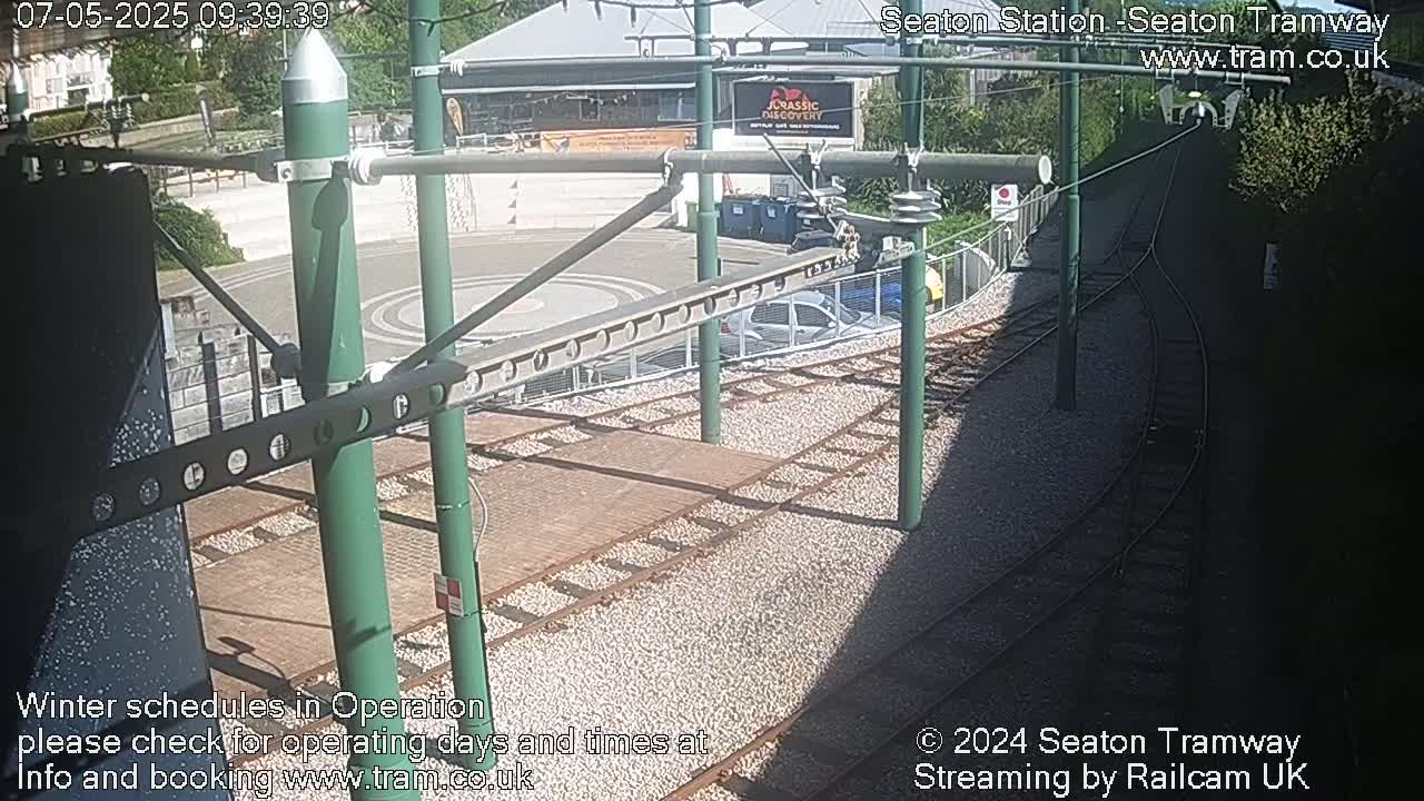 A sunny outdoor view of tram tracks and a station platform, with several cars parked nearby.