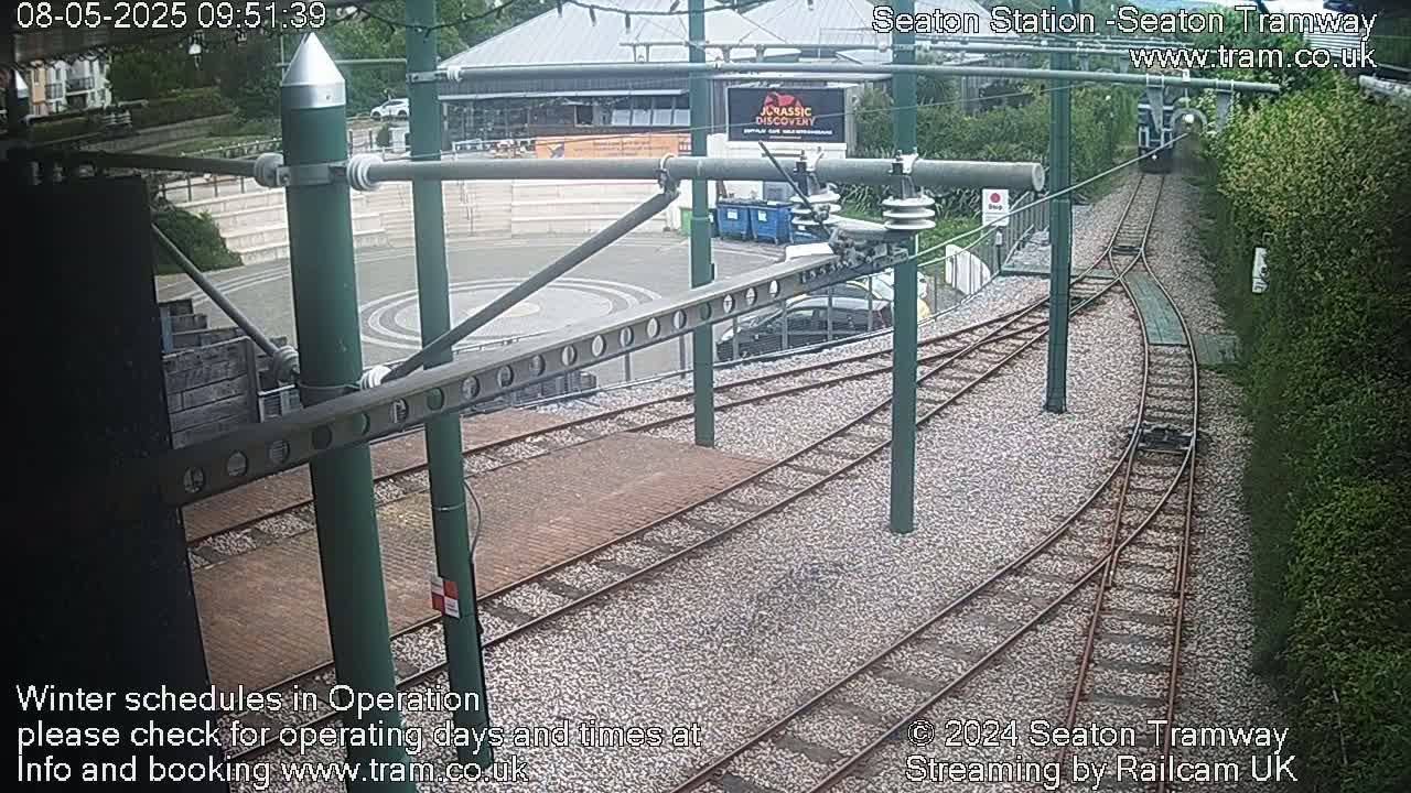Tram tracks curve around a station platform, with a building and greenery visible in the background under an overcast sky.
