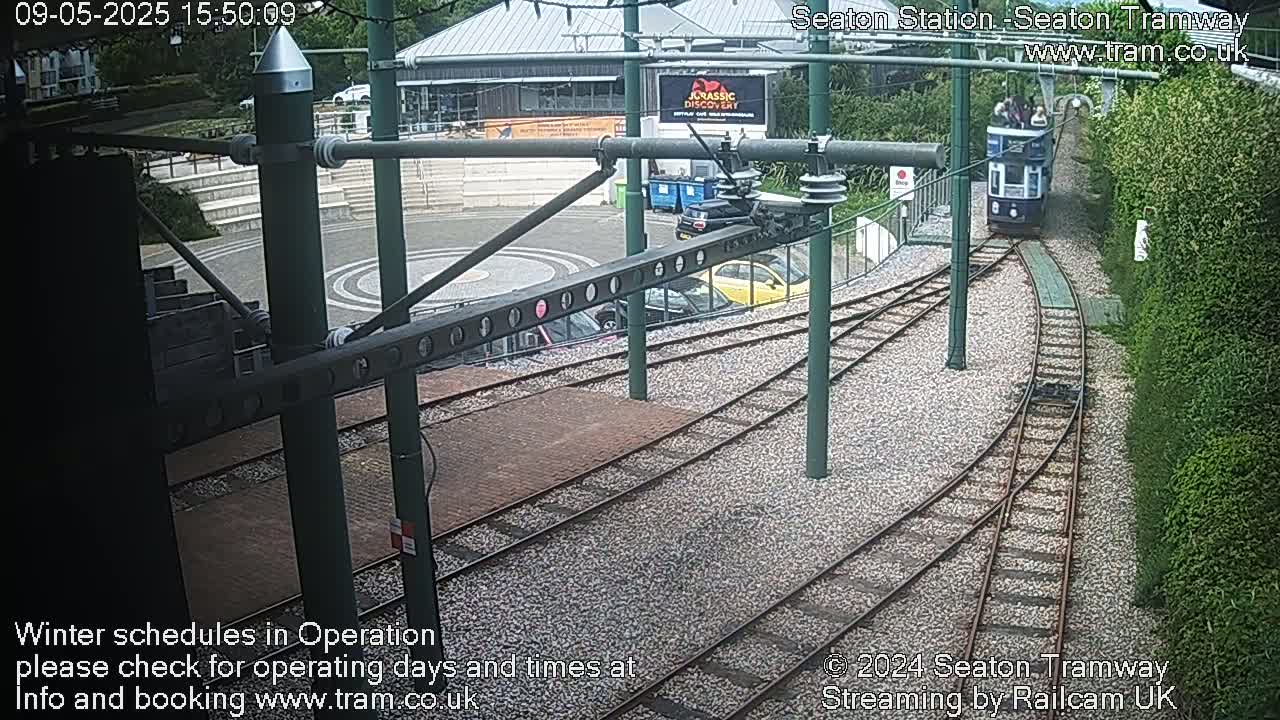 A tram approaches a station, curving onto a set of tracks that intersect another set of tracks, all surrounded by green shrubbery and a stone-paved area.