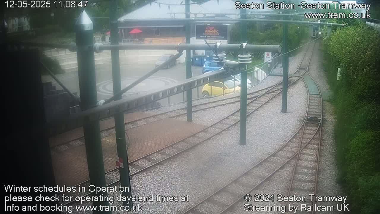 A slightly overcast view of a train station with several cars parked nearby and tram tracks extending into the distance.