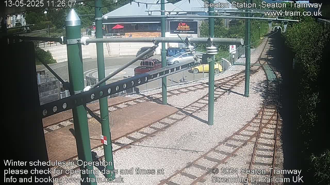A sunny overhead view shows a tram station with several cars parked nearby and train tracks leading away from the station.