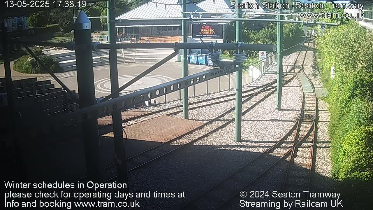 A sunny outdoor view of railway tracks curving around a paved area with seating and a building in the background.