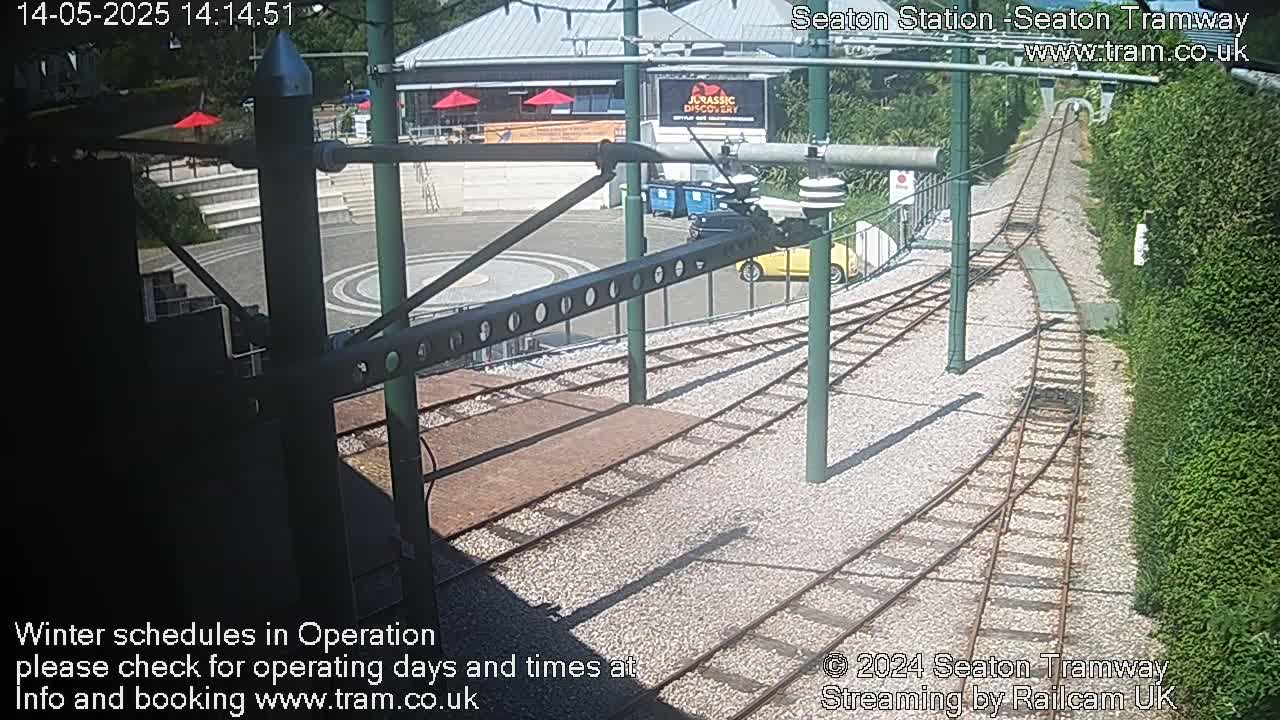 A sunny outdoor scene shows a tram station with train tracks curving away from a small plaza, including a building and some cars.