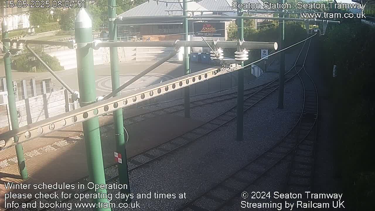 Tram tracks curve through a sunny station area with overhead electrical lines and supporting structures.