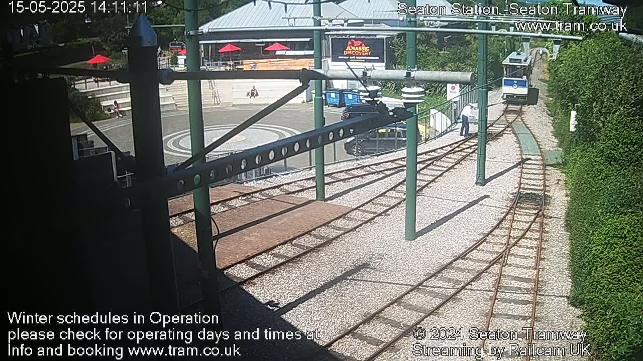 A tram approaches a station on a sunny day, with a person standing nearby and cars parked in the background.