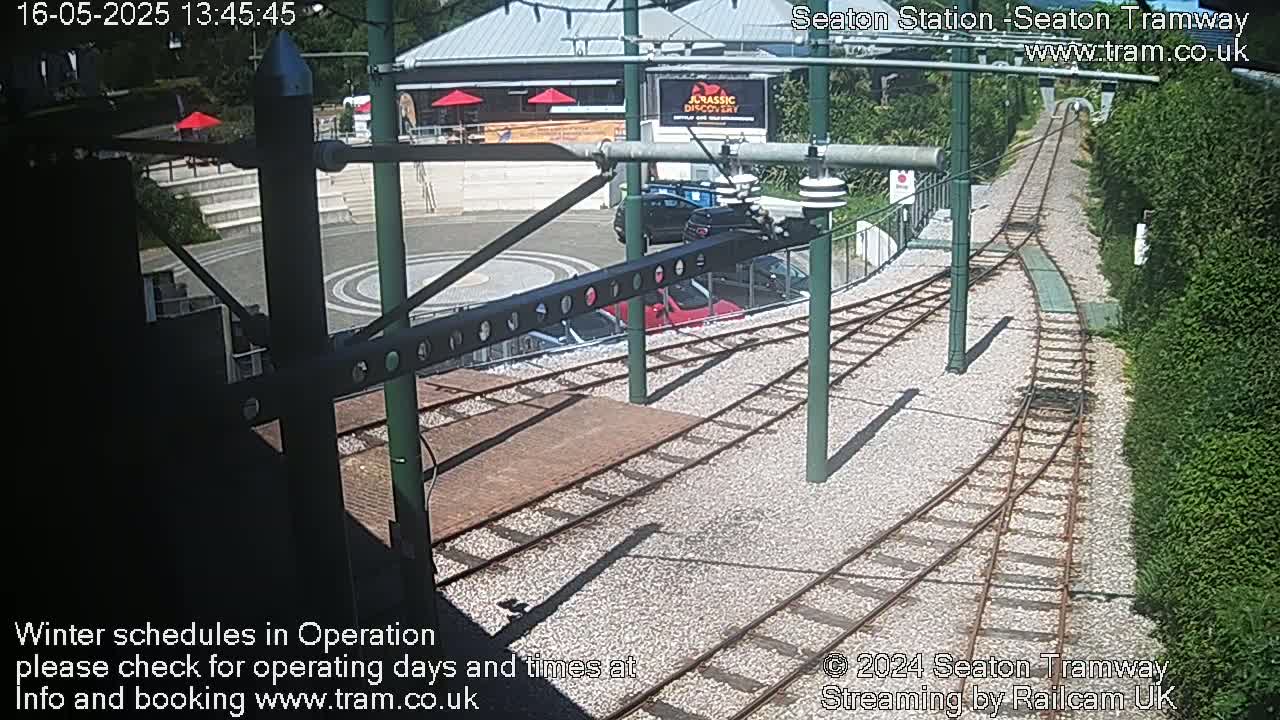 A sunny view of a tramway station with train tracks, a building, and parked cars.