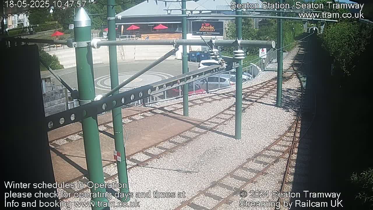 A sunny view of a tram station with train tracks and several parked cars, some green foliage in the background.