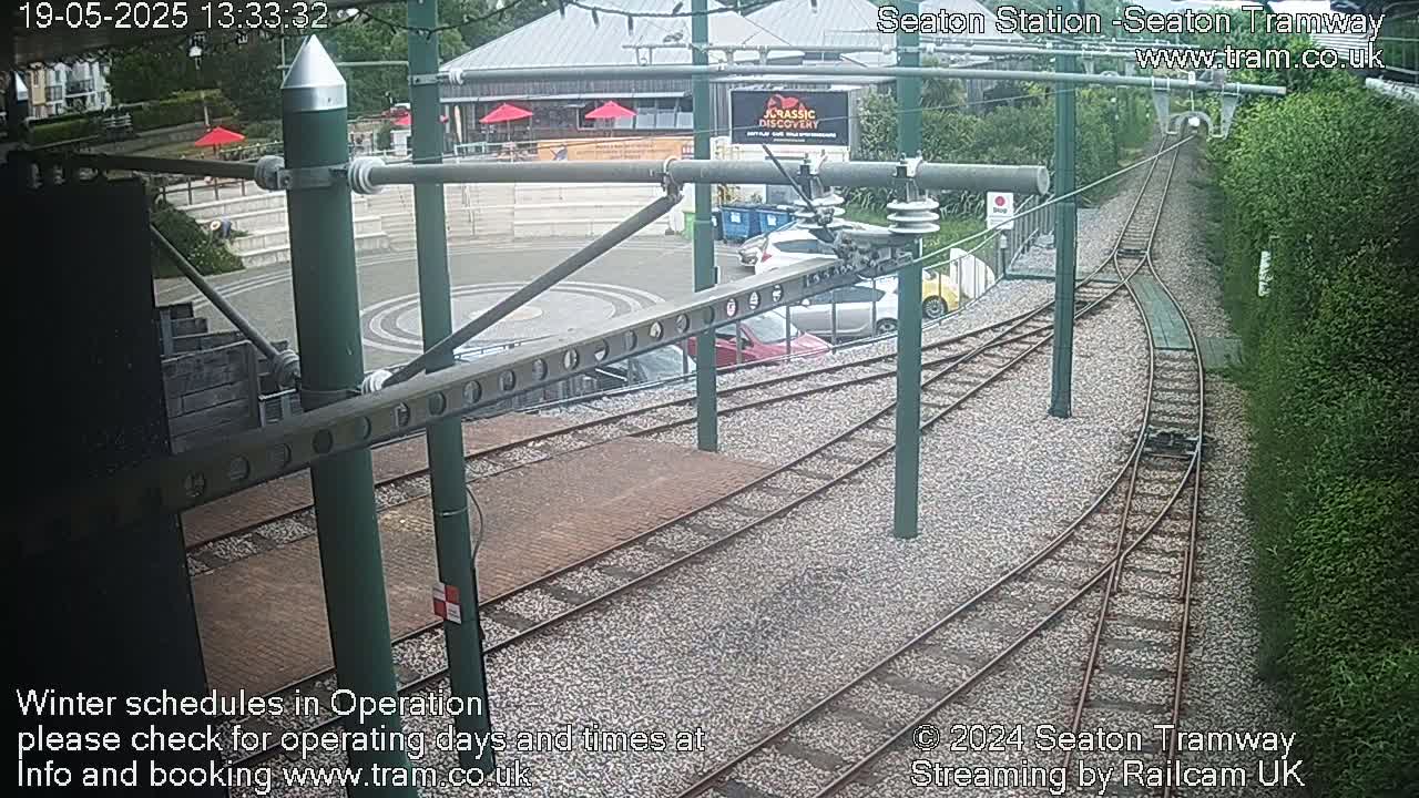 A high-angle view shows a tram station with several cars parked nearby, railway tracks, and green hedges, under overcast skies.