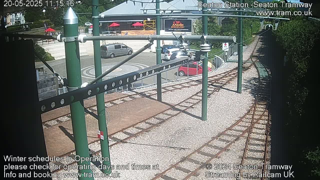 A sunny day at a tram station shows train tracks, a roadway with several cars, and a building with outdoor seating.