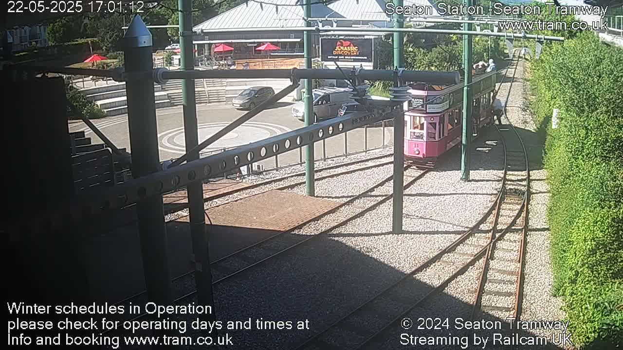 A pink tram is stopped at a station on a sunny day, with cars parked nearby and a grassy embankment in the background.