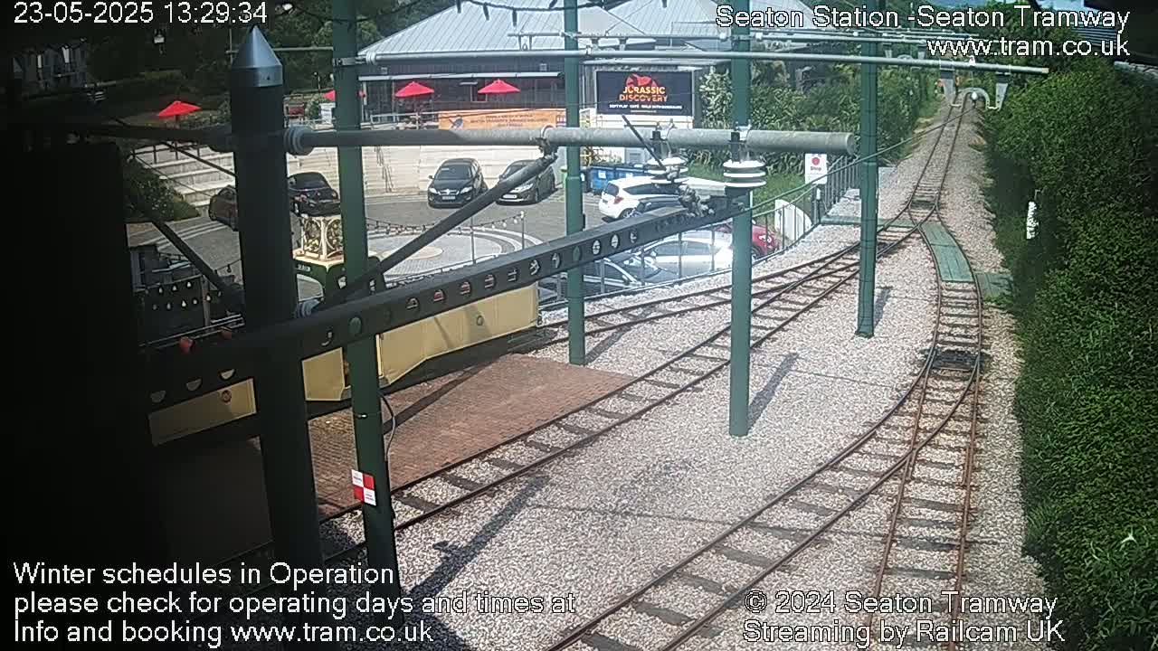 A high-angle view shows a railway station on a sunny day, with several cars parked nearby and train tracks curving away from the camera.