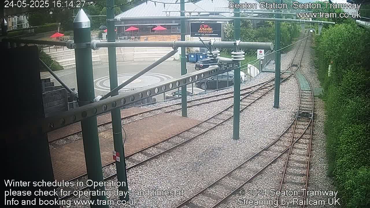 An overhead view shows a tram station with curving tracks, several parked cars, and lush green vegetation under overcast skies.