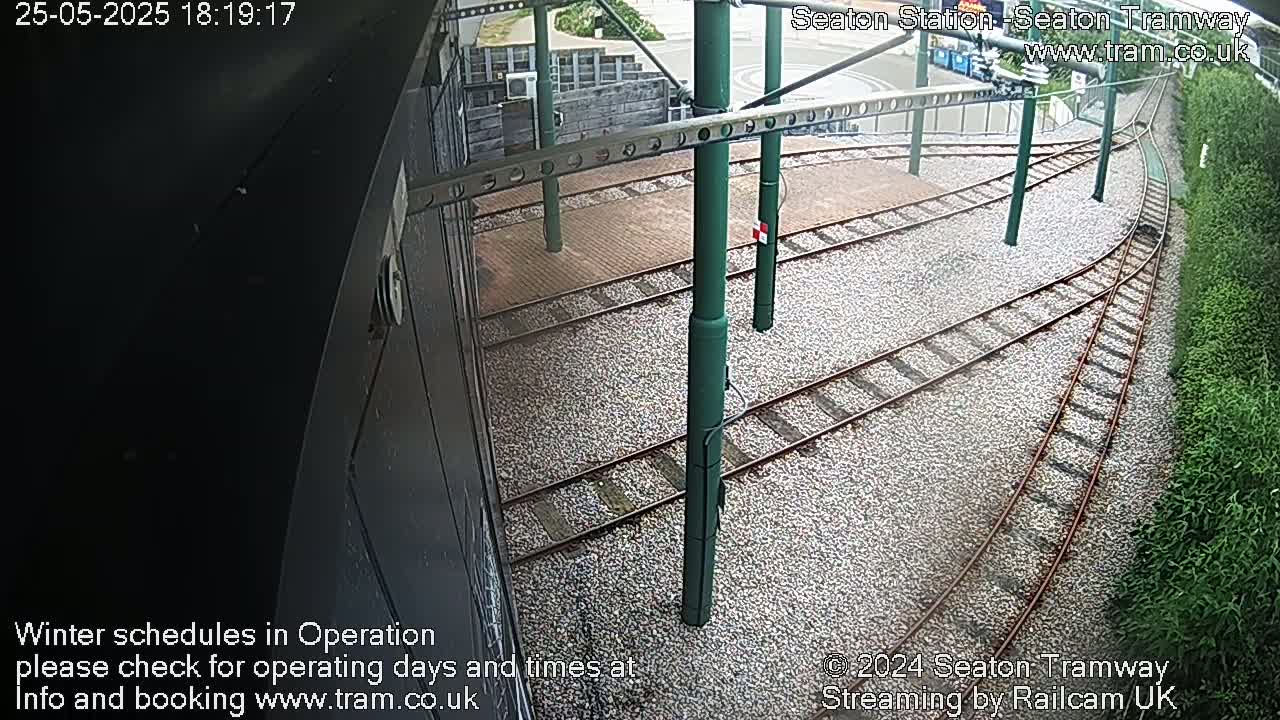 A high-angle view shows a section of tram tracks curving to the right, surrounded by gravel and greenery under an overcast sky.