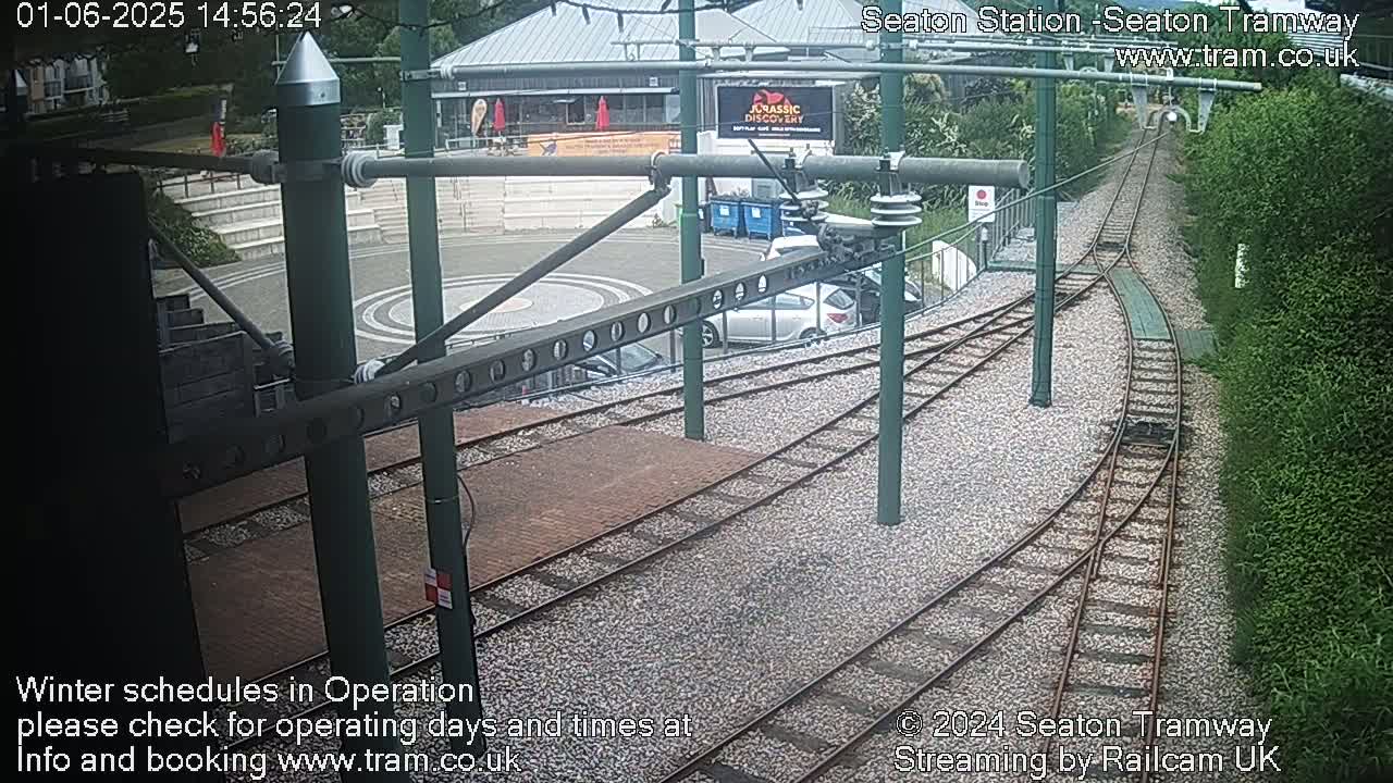 A high-angle view shows a tram station with train tracks curving away, surrounded by landscaping and a building.