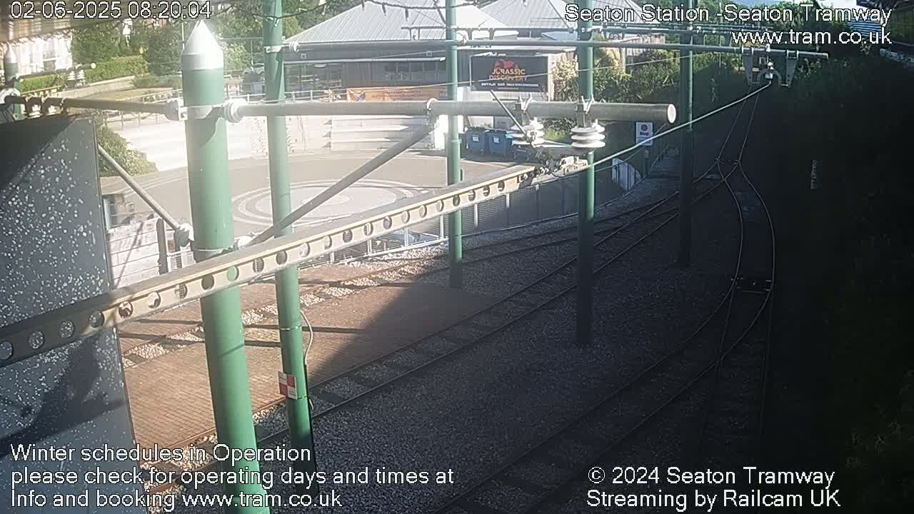 A sunny view of a tram station's track and overhead lines, with a building and some greenery in the background.