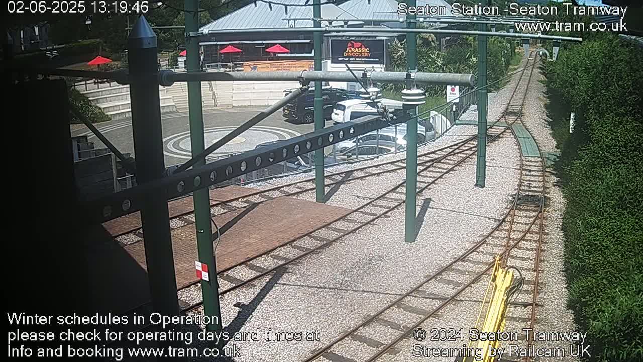 A sunny outdoor scene shows a tram station with train tracks curving into a parking area next to a building.