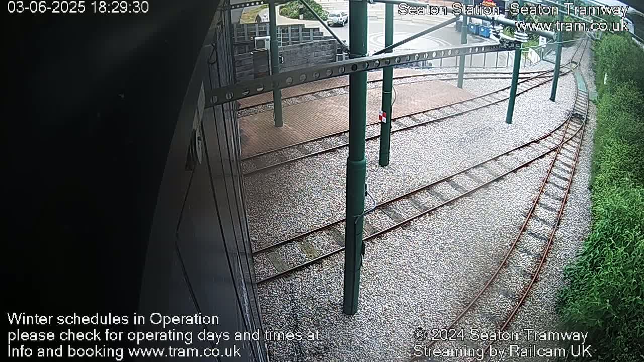 A high-angle view of tram tracks curving through a gravel area next to green vegetation, under overcast conditions.