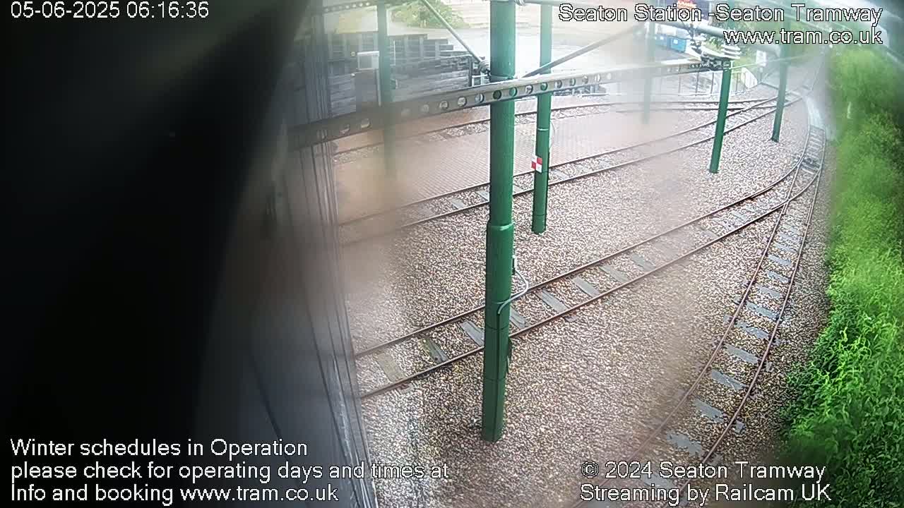 A slightly blurry view of tram tracks and green support poles on a rainy day, with vegetation visible in the lower right corner.