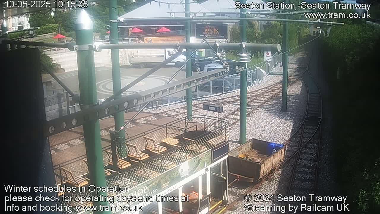 A tram sits at a station on a sunny day next to railway tracks that curve around a paved area with some parked cars and buildings.