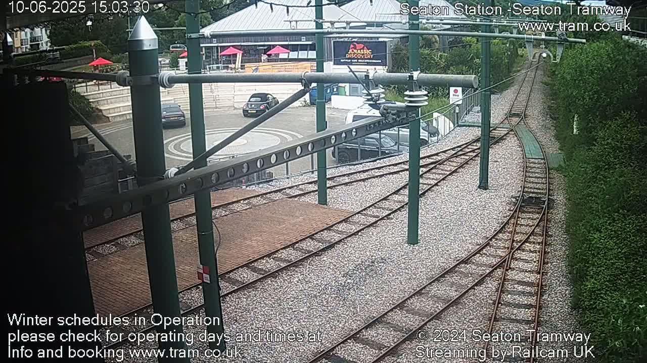 Tram tracks curve through a station area with parked cars and a building visible in the background on a partly sunny day.