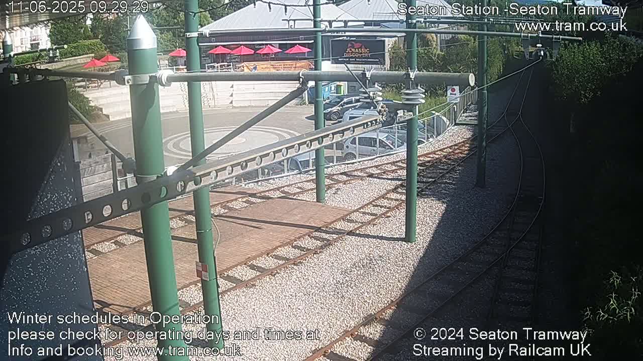 A sunny view of tram tracks and a station platform, with several cars parked nearby and overhead electrical lines.