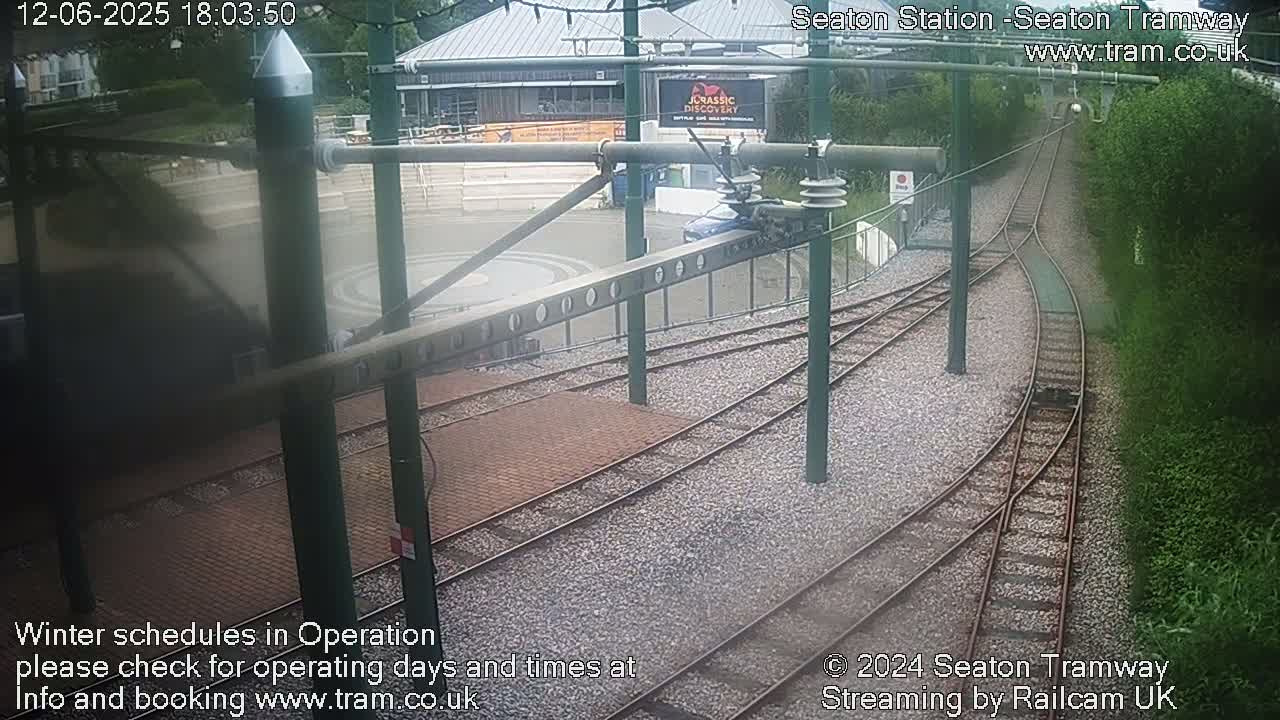 A high-angle view of a tram station's rail tracks and turning area, with a building and lush greenery visible in the background, on a seemingly overcast day.