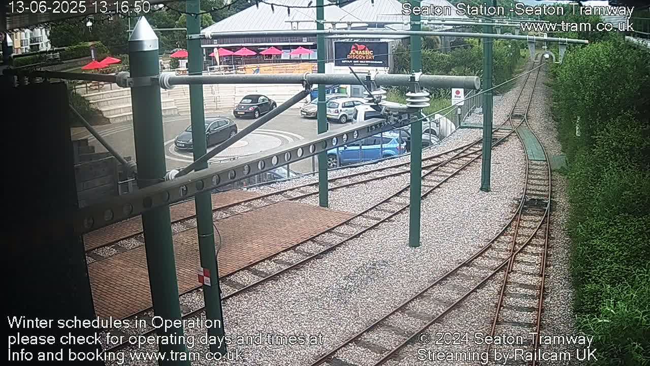 A high-angle view of a tram station with several cars parked nearby and train tracks curving through a gravel area, bordered by greenery.