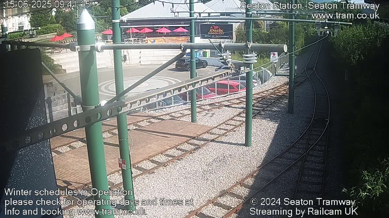 A high-angle view of a tram station on a sunny day, showing train tracks, parked cars, and a building with outdoor seating.
