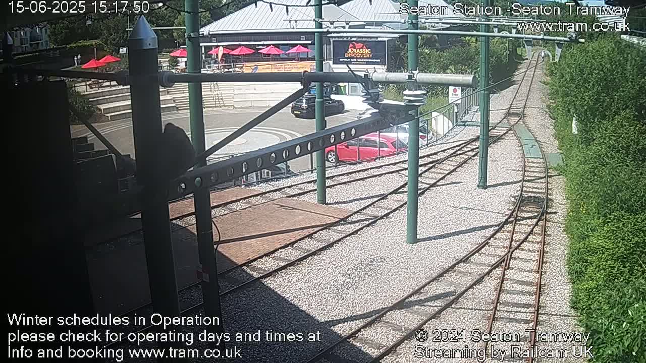 A sunny view of a tram station with a red car and a dark-colored SUV parked nearby, showing railway tracks curving away from the station and surrounded by green vegetation.