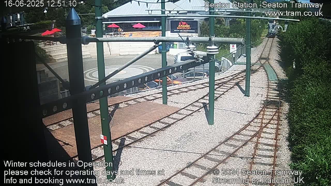 A sunny outdoor view of a train station with railway tracks, a gravel platform, and several parked cars.