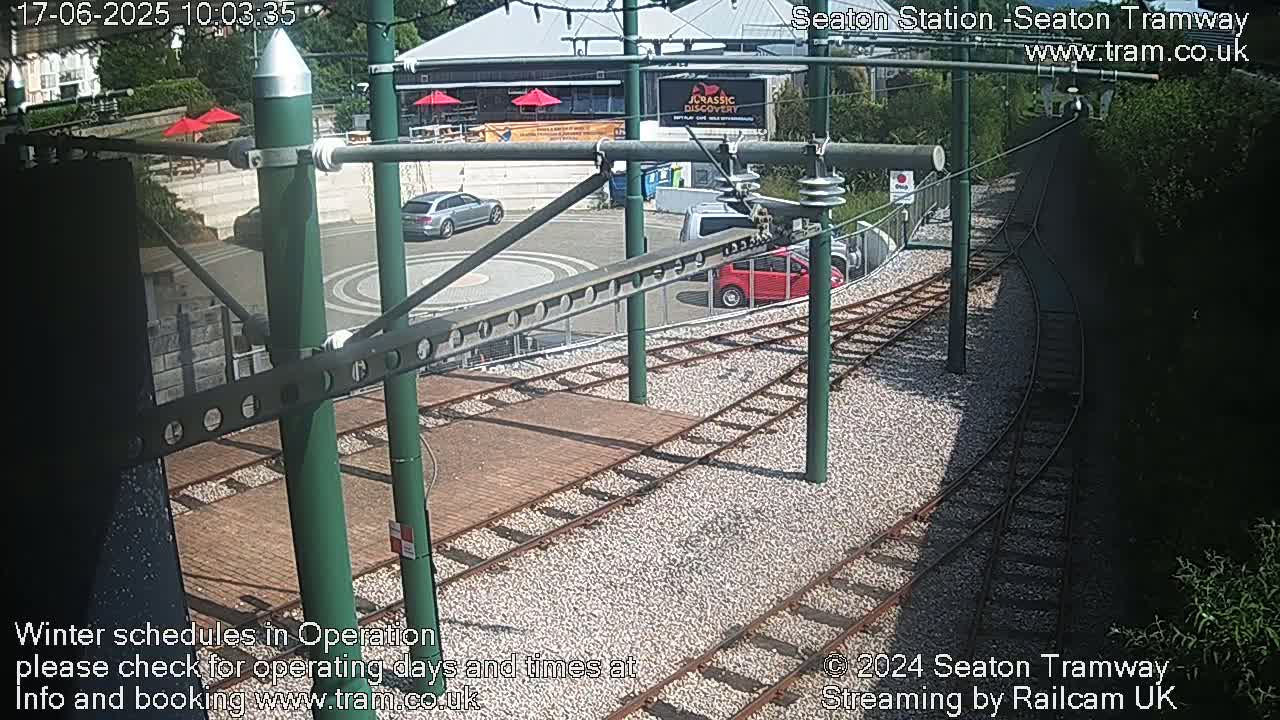 A sunny outdoor scene shows a tram station with several cars parked nearby, tram tracks curving into the distance, and green vegetation bordering the area.