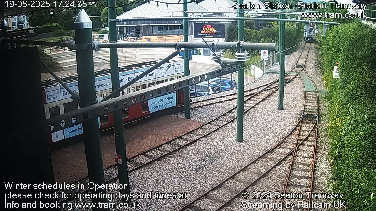 A tram sits at a station on a tram line next to a green hedge, with cars parked nearby under a partly cloudy sky.