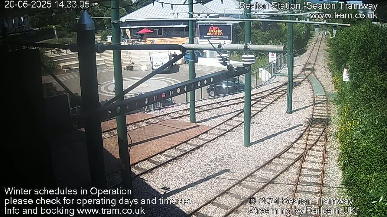 A sunny overhead view of a tram station with tracks curving away from a turning circle, next to a building and green landscaping.